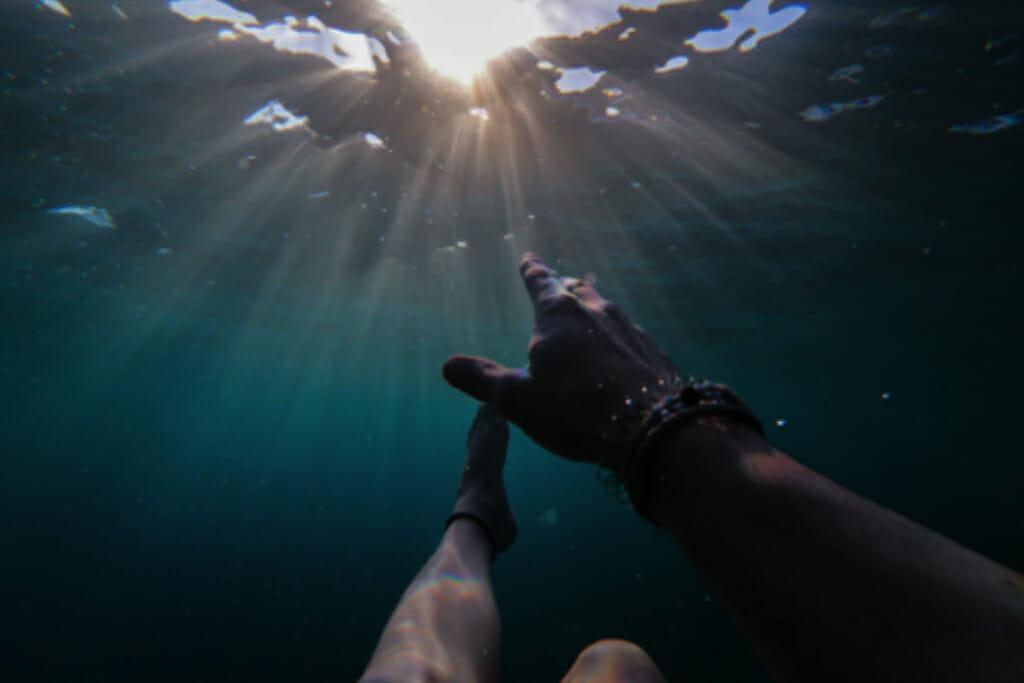 A first-person photograph of a person submerged under water, reaching up for the surface — a metaphor for substance use disorder.