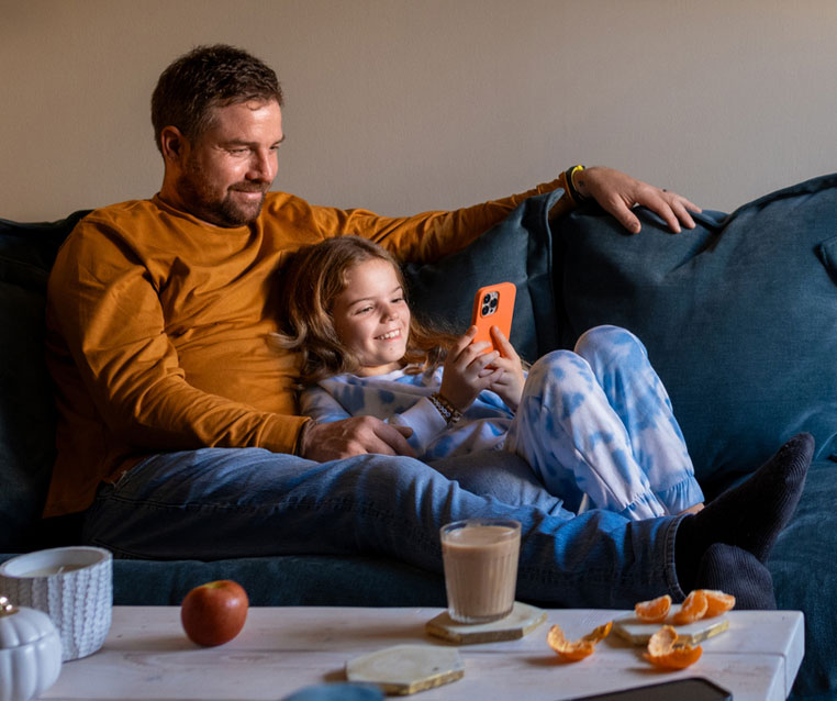 A father and daughter sit together on a sofa, both engaged with a smartphone, sharing a moment of connection.