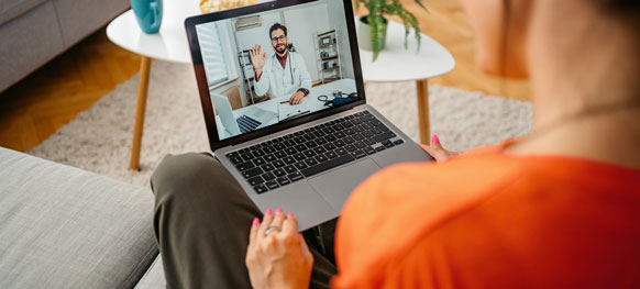 A woman sits on a couch, using a laptop with a man visible on the screen, engaged in a virtual conversation.